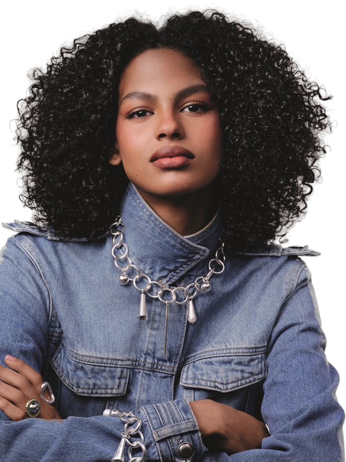 Jovem de cabelo cacheado, com jaqueta jeans e colar prateado, de braços cruzados